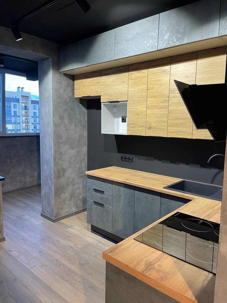 Nowoczesna kuchnia z drewnianymi i szarymi szafkami, czarnym sufitem z oświetleniem punktowym, betonową ścianą i widokiem na budynek mieszkalny z balkonu.