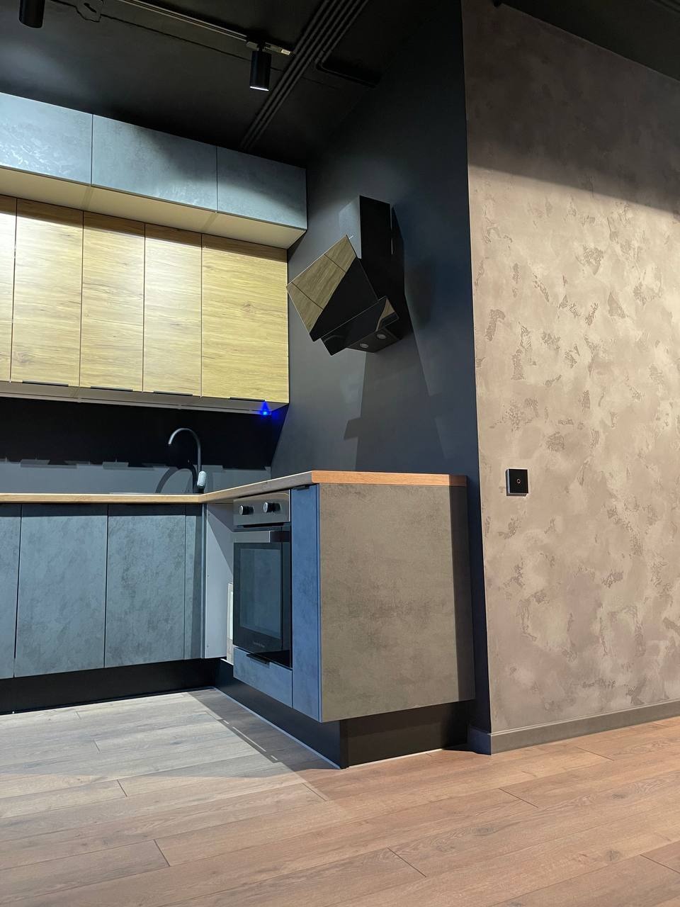 Nowoczesna, minimalistyczna kuchnia z szaro-beżowymi frontami szafek, drewnianym blatem, ciemnoszarą ścianą z fakturą i geometrycznym okapem.