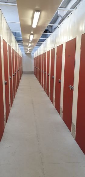Korytarz z rzędem czerwonych boksów do przechowywania, oświetlony szeregiem lamp fluorescencyjnych na suficie.