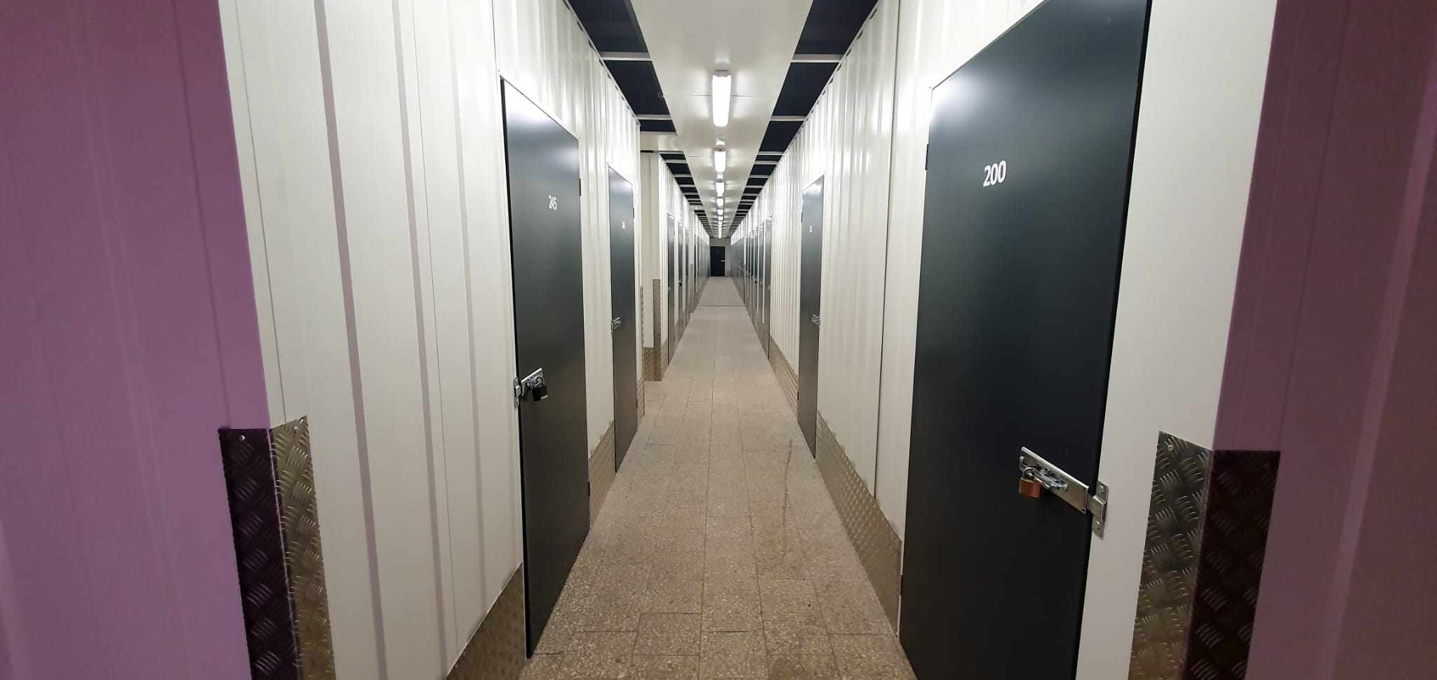 Wnętrze magazynu samoobsługowego z rzędem boksów oznaczonych numerami, widok wzdłuż korytarza z oświetleniem sufitowym i metalowymi zabezpieczeniami ścian.