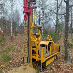 Margeo Grzegorz Markiewicz - Żółto-czerwona wiertnica na gąsienicach w lesie, wiertło z nawierconą ziemią, liście na ziemi, drzewa bez liści w tle.