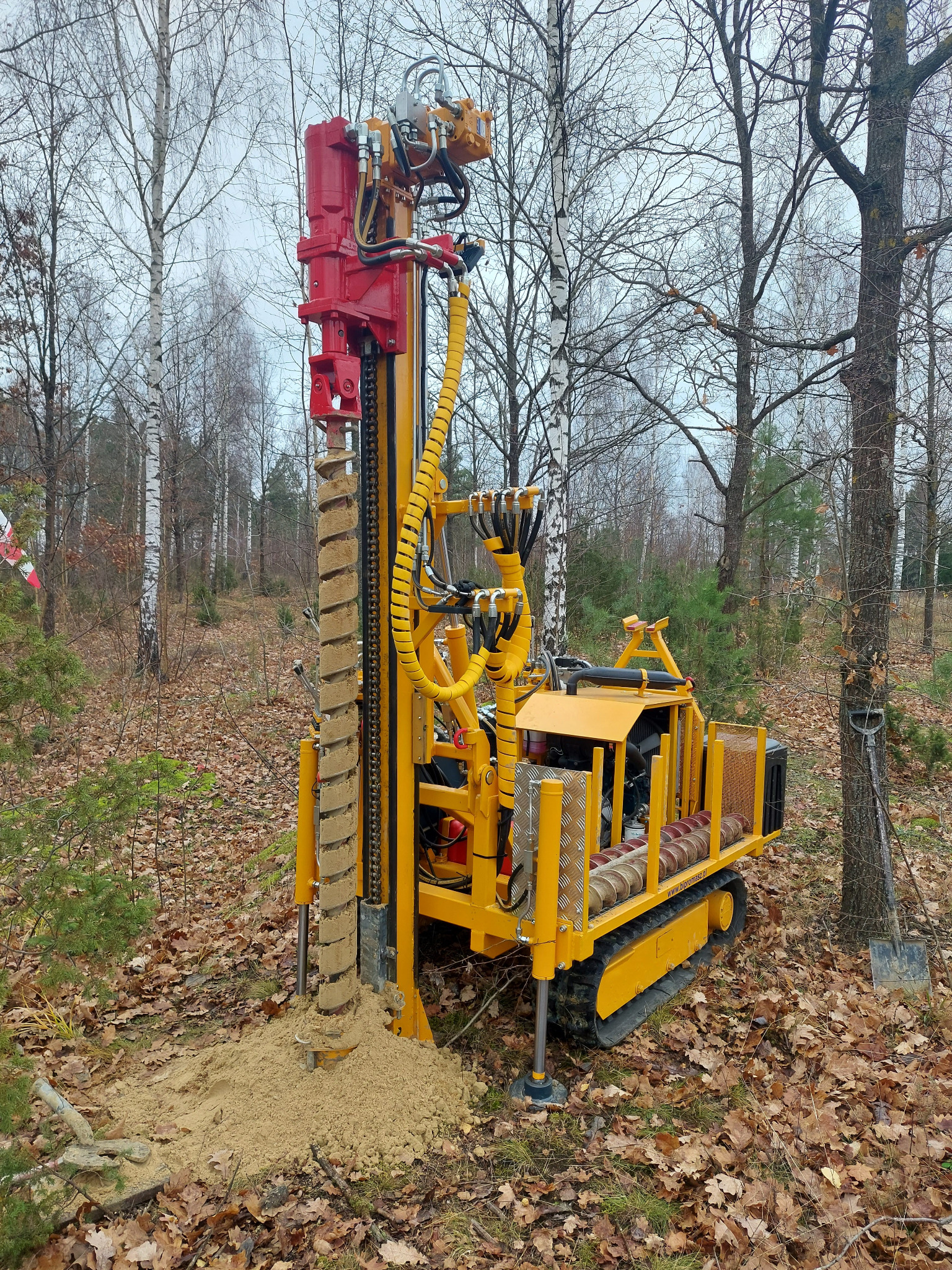 Żółto-czerwona wiertnica na gąsienicach w lesie, wiertło z nawierconą ziemią, liście na ziemi, drzewa bez liści w tle.