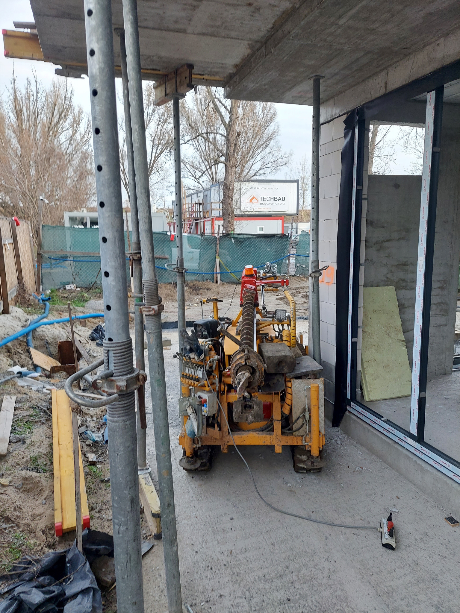 Wiertnica do gruntu na placu budowy, widoczne szalunki i elementy konstrukcyjne budynku w surowym stanie.