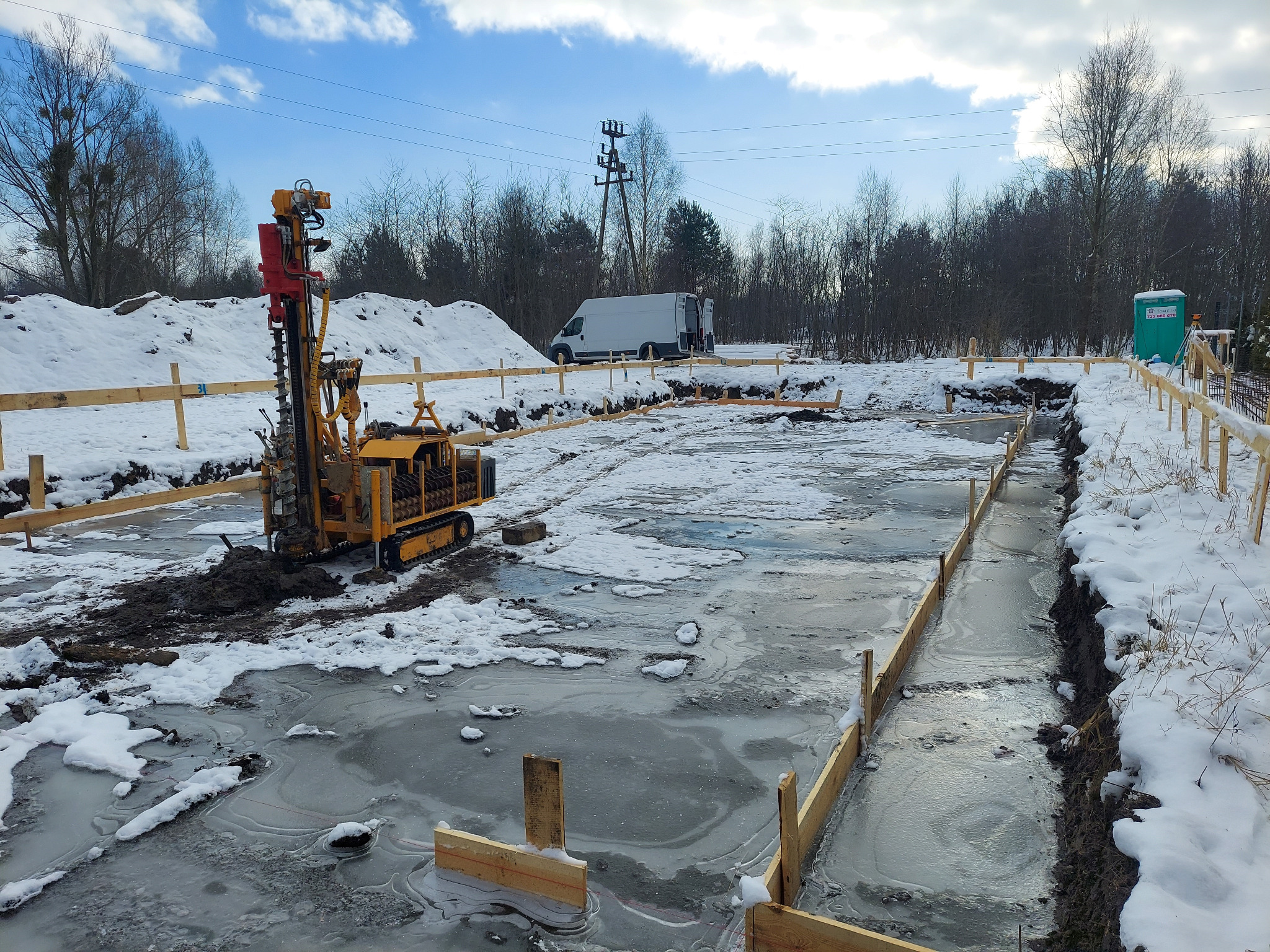 Żółta wiertnica gąsienicowa w zasypanym śniegiem wykopie fundamentowym, z widocznymi deskami szalunkowymi i cienką warstwą lodu na dnie wykopu, w tle biały samochód dostawczy i zasypane śniegiem pole.