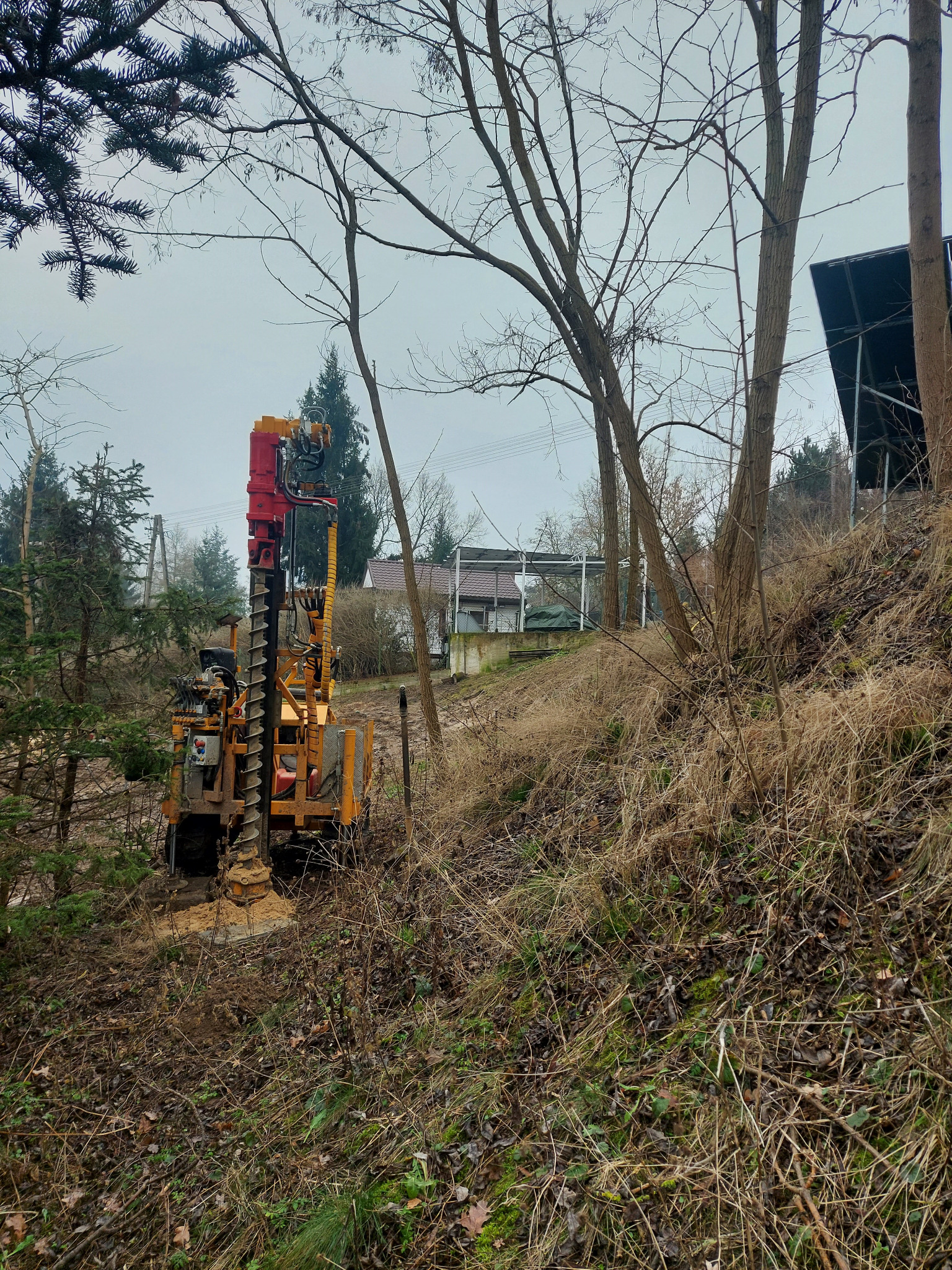 Żółto-czerwona wiertnica geologiczna w trakcie wiercenia otworu w ziemi na zalesionym zboczu, widok z dołu, z domem w tle.