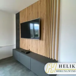Szara szafka RTV z ukrytym systemem przechowywania, zamontowana pod telewizorem na ścianie z drewnianymi panelami dekoracyjnymi i czarnym akcentem, widoczne logo firmy Helik Meble na Wymiar.