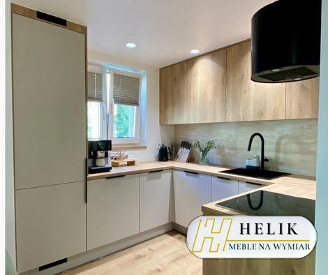 Jasna kuchnia z drewnianymi akcentami: zabudowa z szarymi frontami, drewniane szafki wiszące, czarny okap i blat. Minimalistyczny design z oknem w tle.