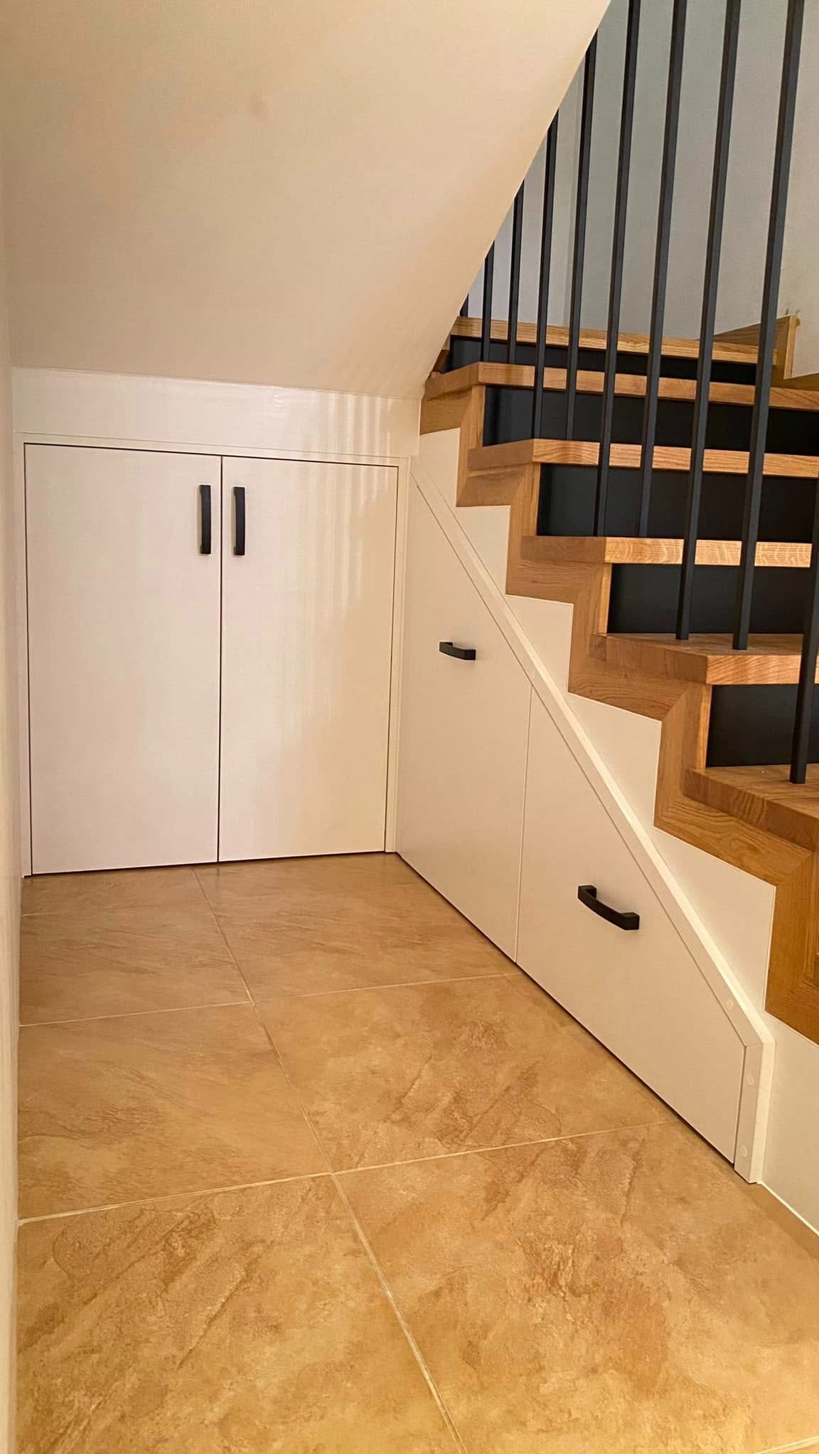 Białe szafy wnękowe pod schodami z czarnymi uchwytami, drewniane schody z czarnymi balustradami, brązowe płytki podłogowe. Funkcjonalne zagospodarowanie przestrzeni.