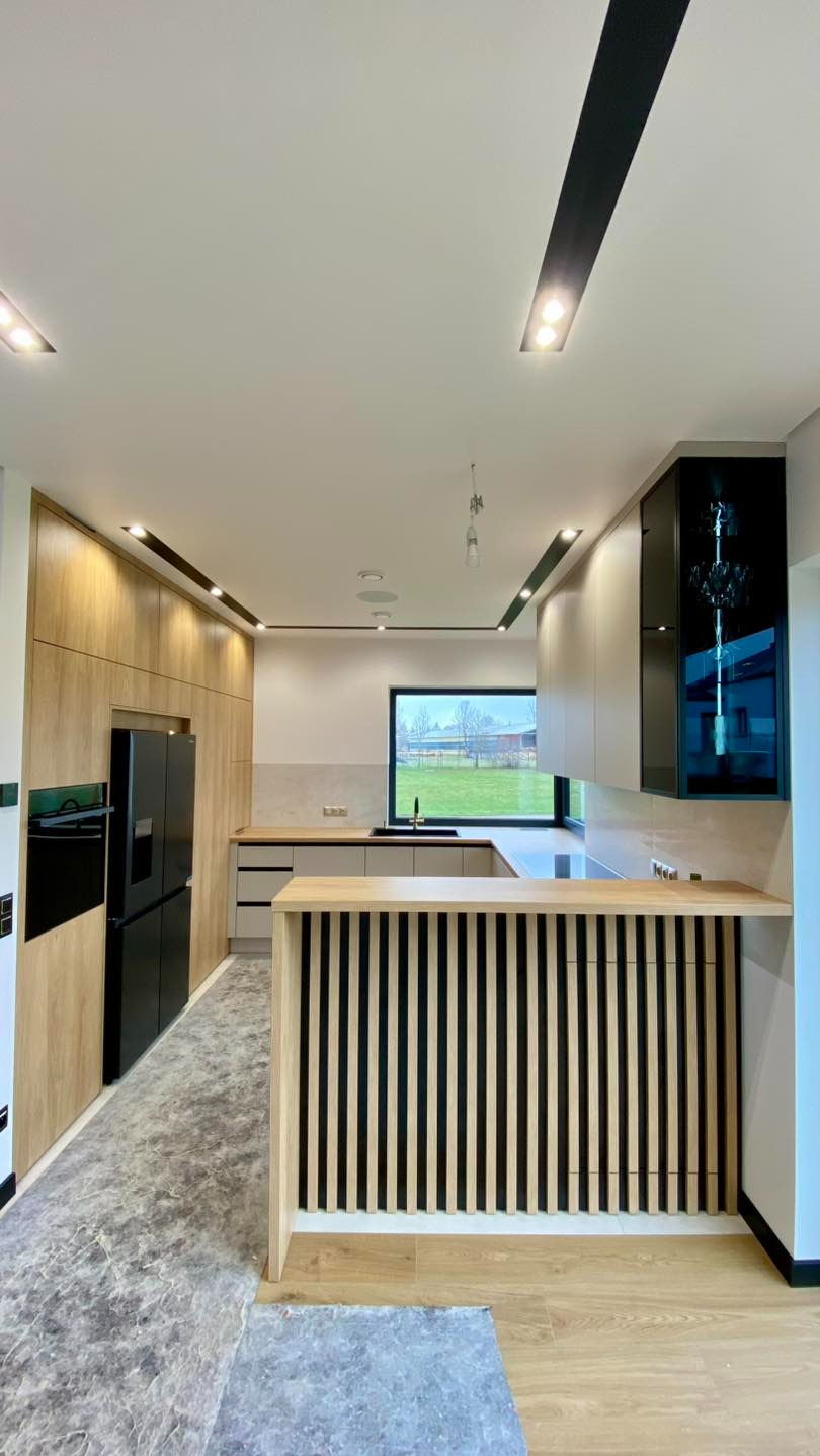 Nowoczesna kuchnia z drewnianą zabudową, czarnym sprzętem AGD i oryginalnym barem z pionowych listewek. Widok na zielony krajobraz przez okno.