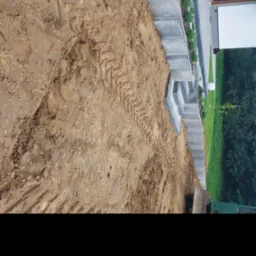 Przygotowany teren z widocznymi śladami opon maszyny budowlanej, obok schody z kostki brukowej prowadzące do nowoczesnego domu z ogrodem.