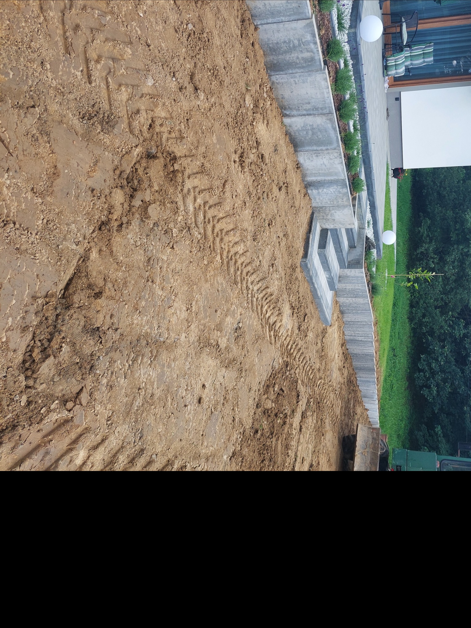 Przygotowany teren z widocznymi śladami opon maszyny budowlanej, obok schody z kostki brukowej prowadzące do nowoczesnego domu z ogrodem.