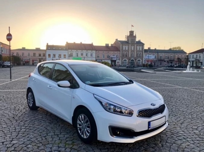Biały samochód marki Kia zaparkowany na brukowanym placu miejskim o wschodzie słońca, z widocznymi zabytkowymi budynkami w tle.