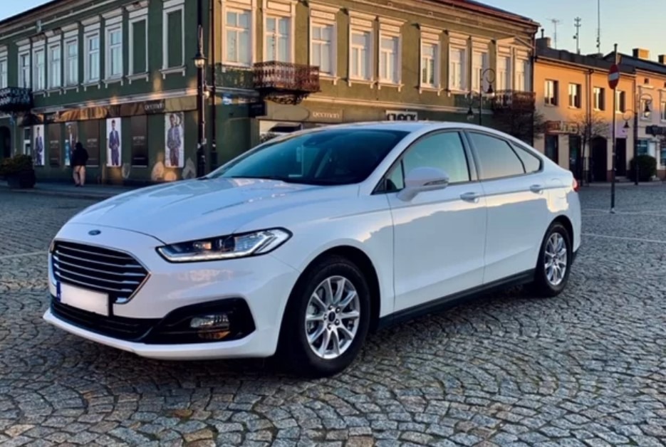 Biały sedan na brukowanym placu miejskim, budynek w tle z zielonymi elementami architektonicznymi i reklamami w oknach.