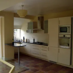 Jasna kuchnia z beżowymi frontami, czarnym blatem, piekarnikiem i mikrofalówką w zabudowie oraz barkiem z pojedynczą nogą, widok na ogród przez otwarte drzwi balkonowe.