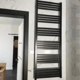 Czarny, drabinkowy grzejnik łazienkowy zamontowany na ścianie wyłożonej białymi płytkami imitującymi marmur, widok z perspektywy drzwi z czarną ościeżnicą do innego pomieszczenia z ciemną podłogą.