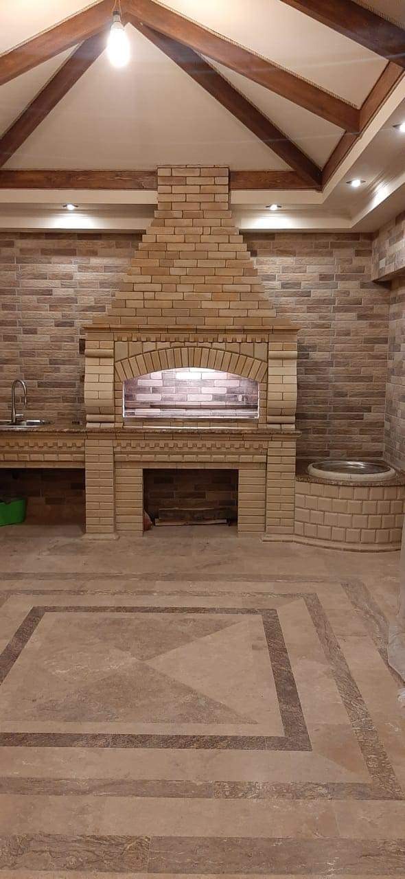 Murowany piec w pomieszczeniu z wykończonymi ścianami z płytek imitujących kamień, drewnianą konstrukcją sufitu i dekoracyjną podłogą z płytek.