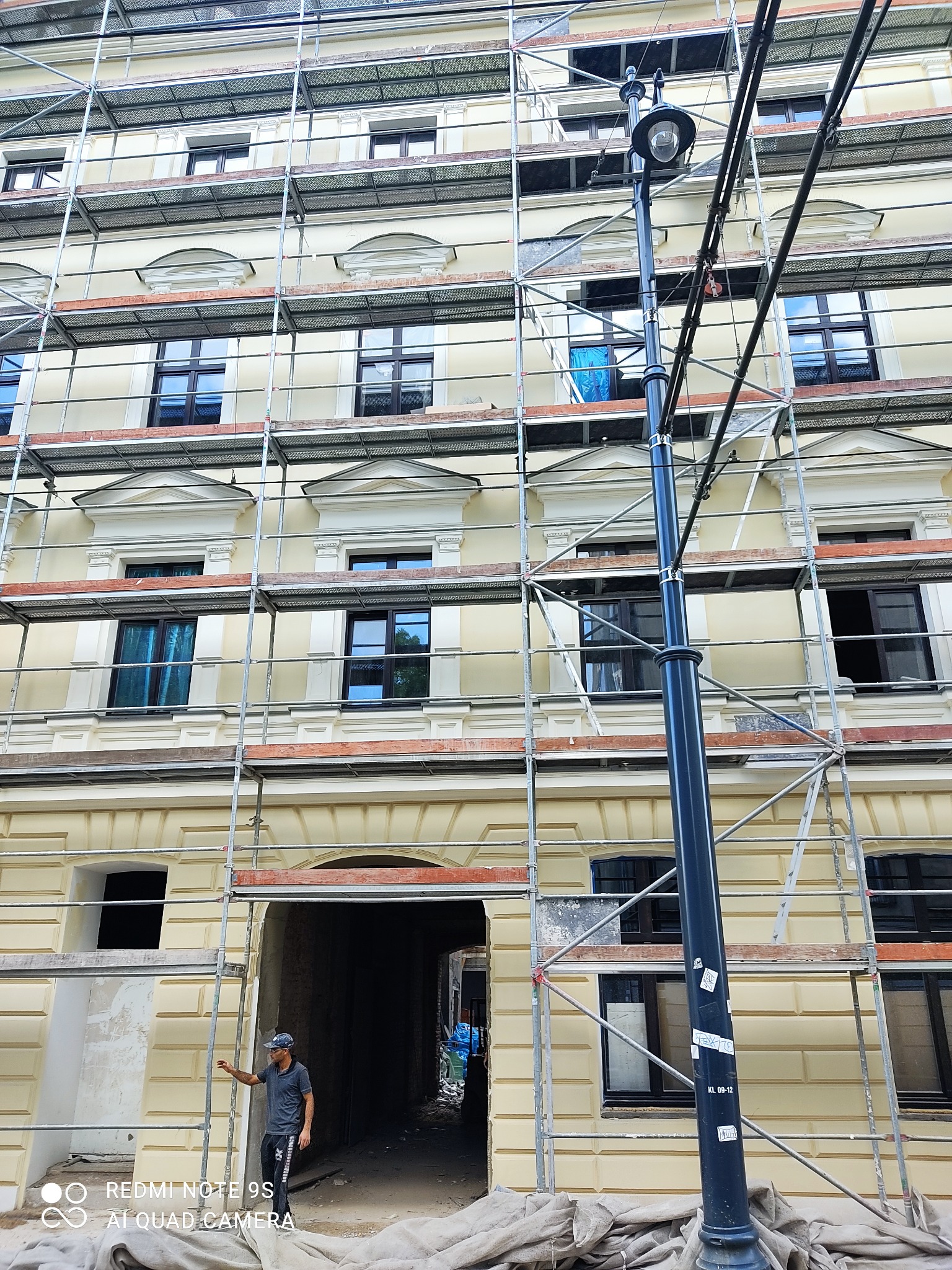 Elewacja budynku w trakcie prac remontowych, widoczne rusztowania i fragment odnowionej fasady z oknami, mężczyzna w tle wskazujący na postęp prac.