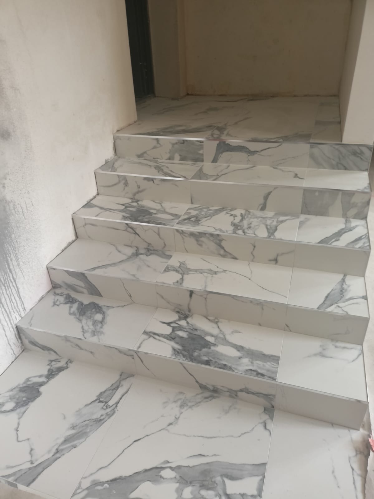 Schody wyłożone białymi płytkami imitującymi marmur, widoczne fugi i aluminiowe listwy wykończeniowe na krawędziach stopni, ściana z widocznymi śladami niedokończonych prac.