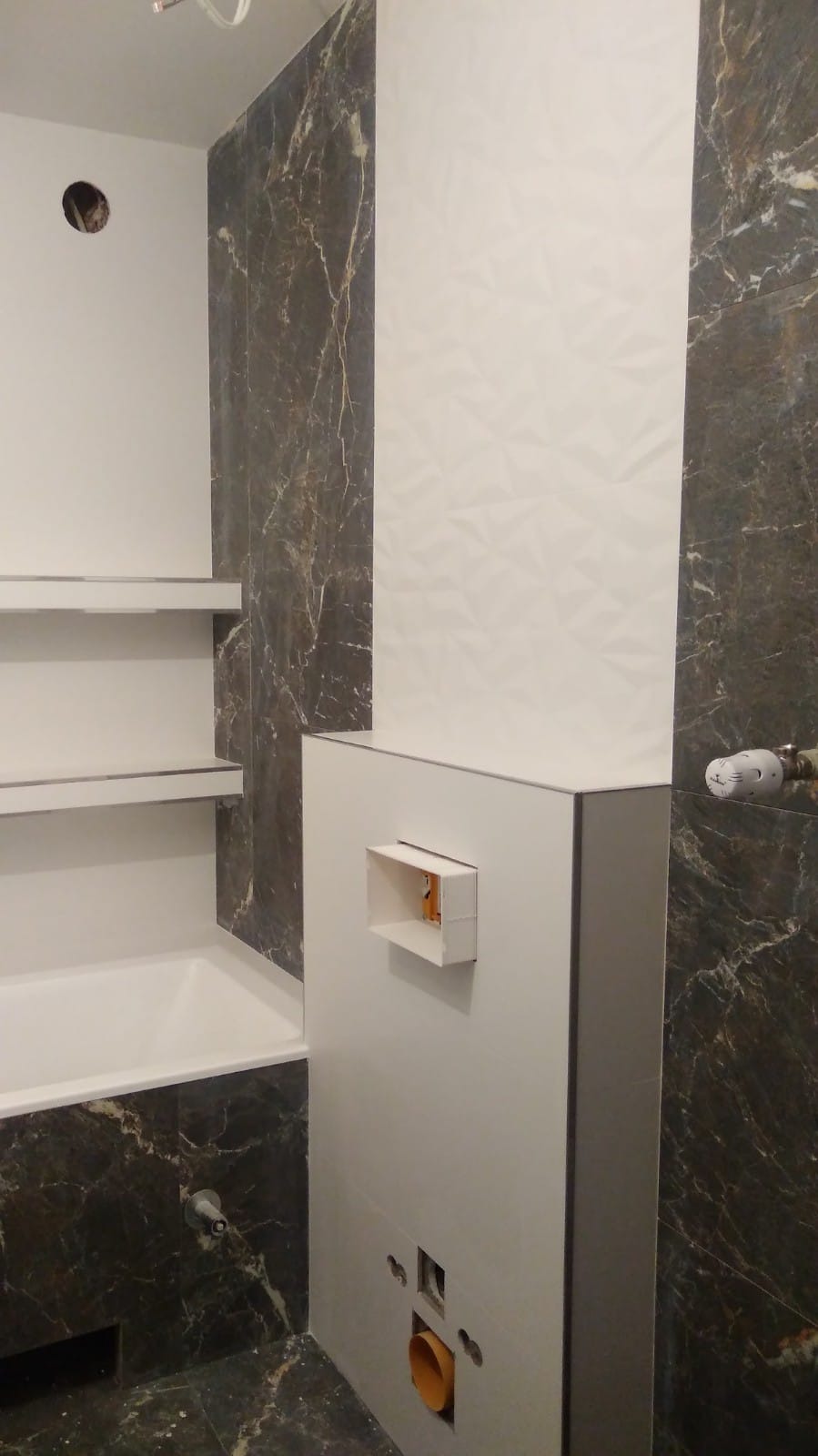 Fragment łazienki w trakcie wykańczania: ściana z płytek imitujących marmur, biała ściana z trójwymiarowym wzorem, zabudowa z otworem rewizyjnym i widoczną rurą kanalizacyjną.