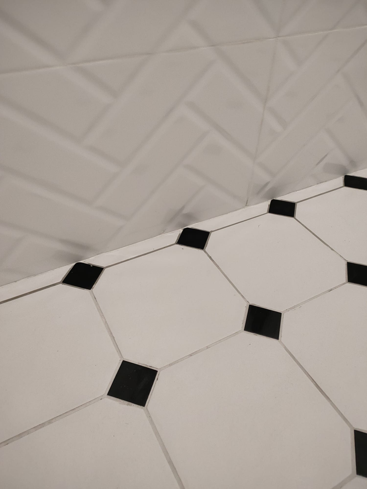 Białe płytki podłogowe z czarnymi akcentami ułożone w romby, przylegające do ściany z białych płytek ułożonych w jodełkę.