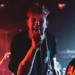 Młody mężczyzna z tatuażem na ramieniu śpiewa do mikrofonu na scenie w czerwonym oświetleniu, widoczne bębny w tle.