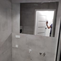 Efekt-Fliz - Wykończona łazienka w Pogórskiej Woli z szarymi płytkami, dużym lustrem, drewnopodobną podłogą i widoczną instalacją wodną.