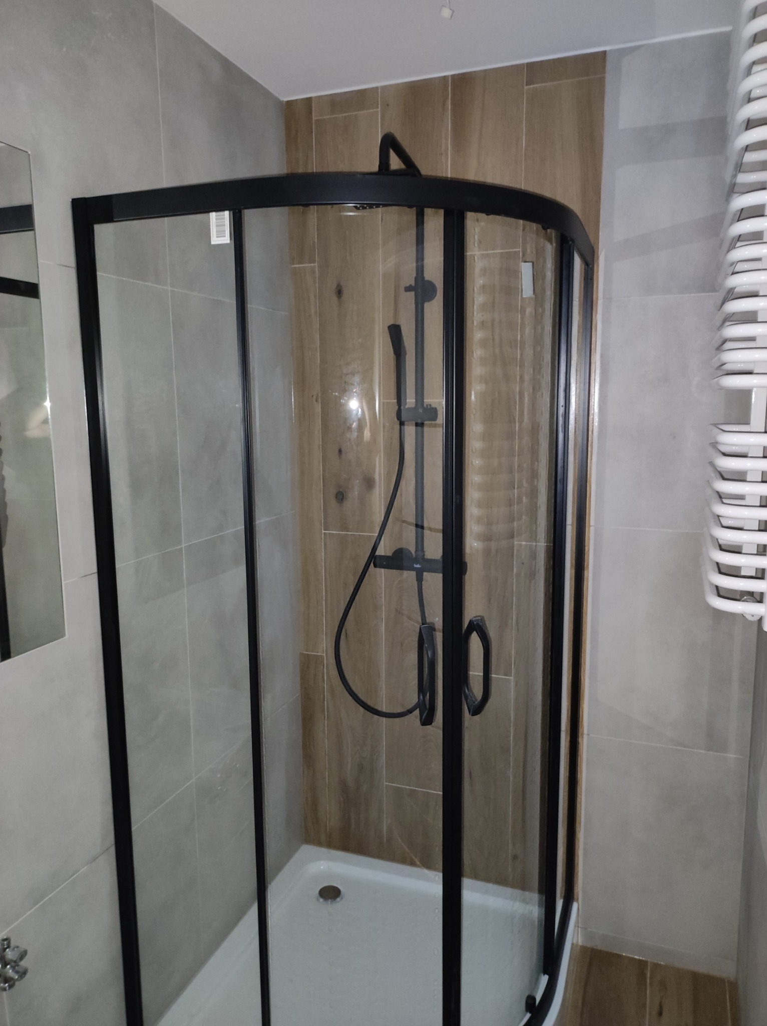 Kabina prysznicowa z czarną ramą, zamontowana w rogu łazienki wyłożonej szarymi i drewnopodobnymi płytkami, widoczny grzejnik drabinkowy.