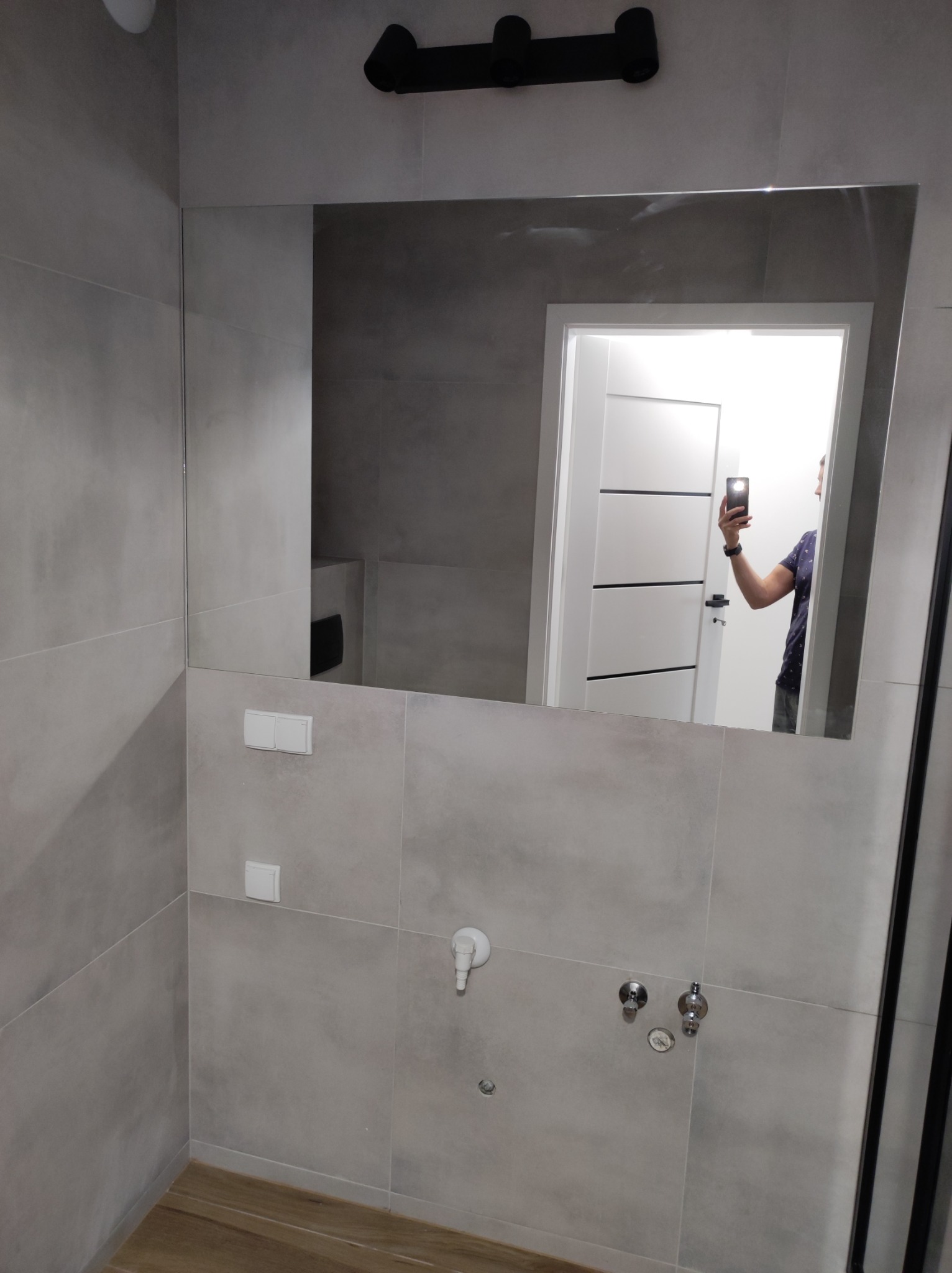 Wykończona łazienka w Pogórskiej Woli z szarymi płytkami, dużym lustrem, drewnopodobną podłogą i widoczną instalacją wodną.