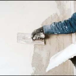 Nakładanie gładzi szpachlą na ścianę, widoczne przejście między surowym murem a świeżo nałożoną warstwą.