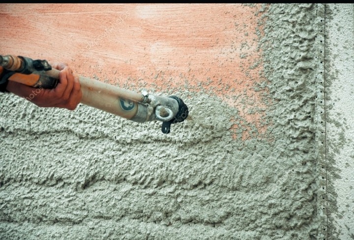 Aplikacja świeżego, szarego tynku na ścianę za pomocą pistoletu natryskowego; widoczny fragment starej, pomarańczowej warstwy spod spodu.