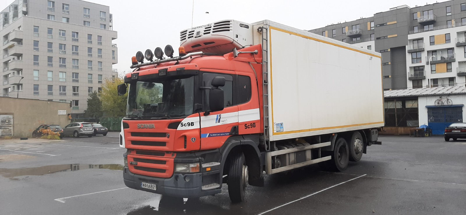 Czerwona ciężarówka Scania z chłodnią Thermo King, zaparkowana na mokrym placu, w tle budynki mieszkalne i garaż z rowerem na dachu.