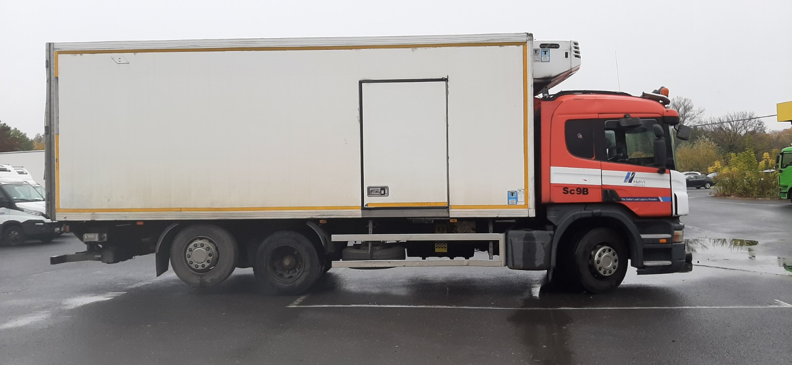 Czerwono-biała ciężarówka chłodnia Scania Sc9B z agregatem chłodniczym na dachu, zaparkowana na mokrym parkingu.