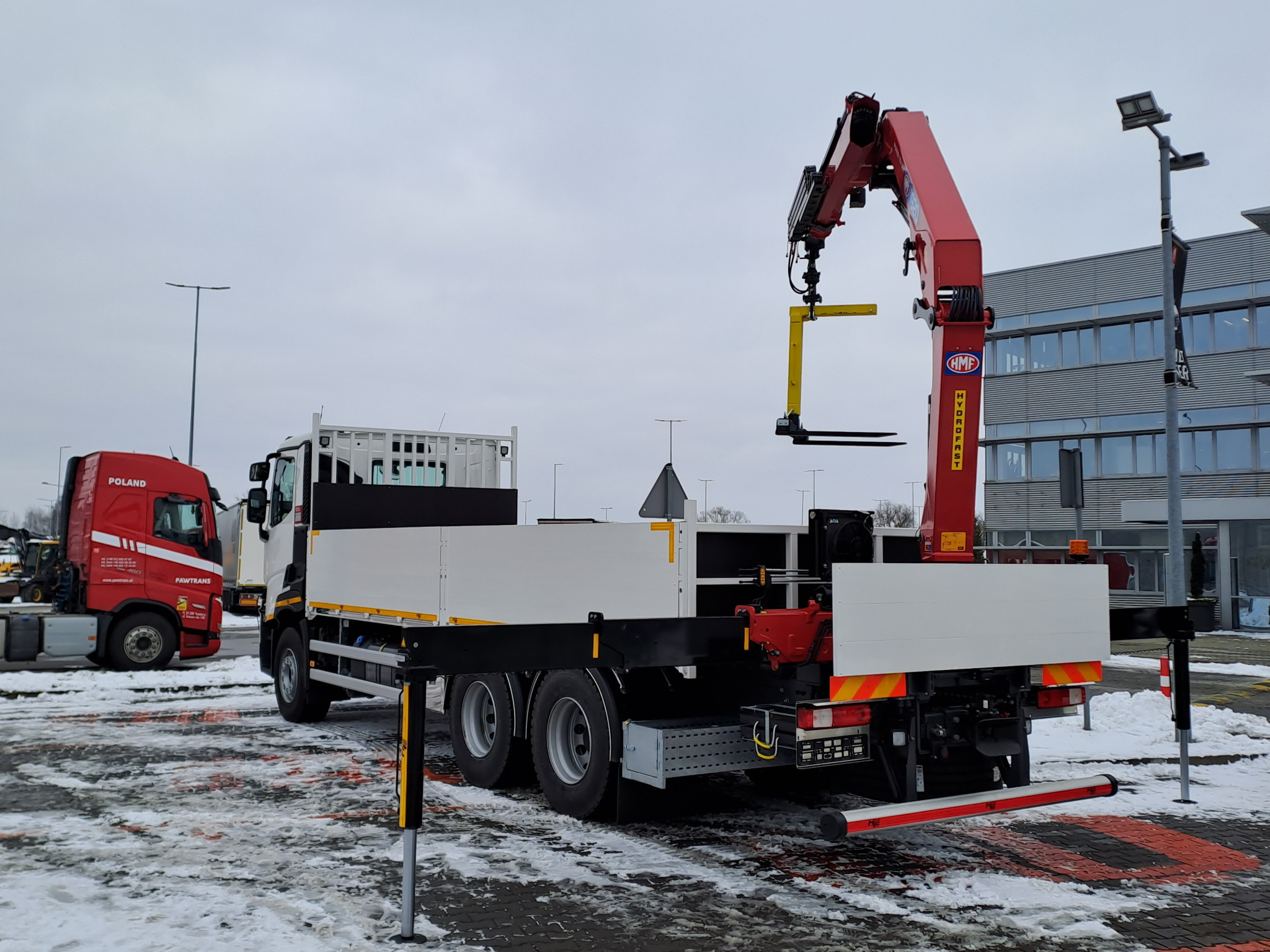 Ciężarówka z zamontowanym czerwonym dźwigiem HMF na parkingu, widoczna tablica rejestracyjna i częściowo ośnieżona nawierzchnia.