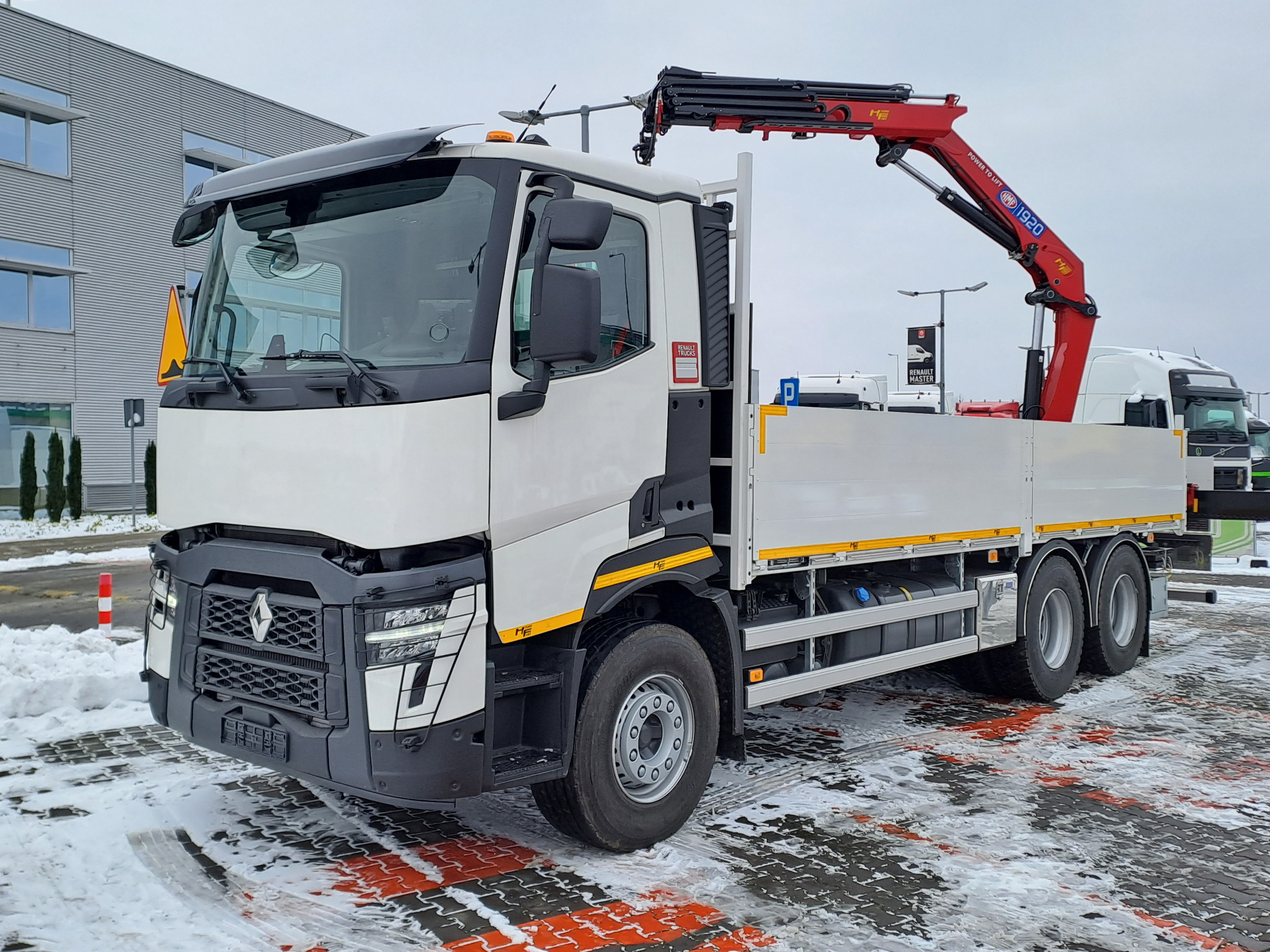 Biała ciężarówka Renault z czerwoną hydrauliką dźwigową HMF 1920, zaparkowana na placu pokrytym śniegiem i kostką brukową z pomarańczowymi pasami.
