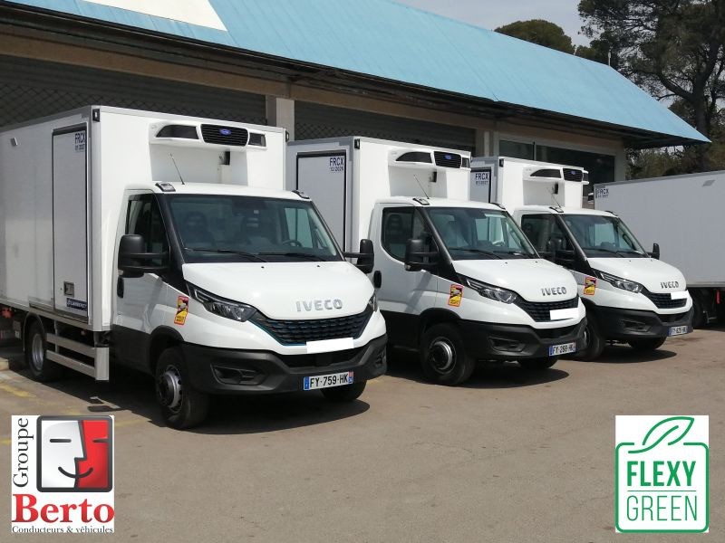 Rząd białych samochodów dostawczych Iveco z chłodniami, zaparkowanych przed budynkiem o niebieskim dachu, z widocznymi logotypami Groupe Berto i Flexy Green.