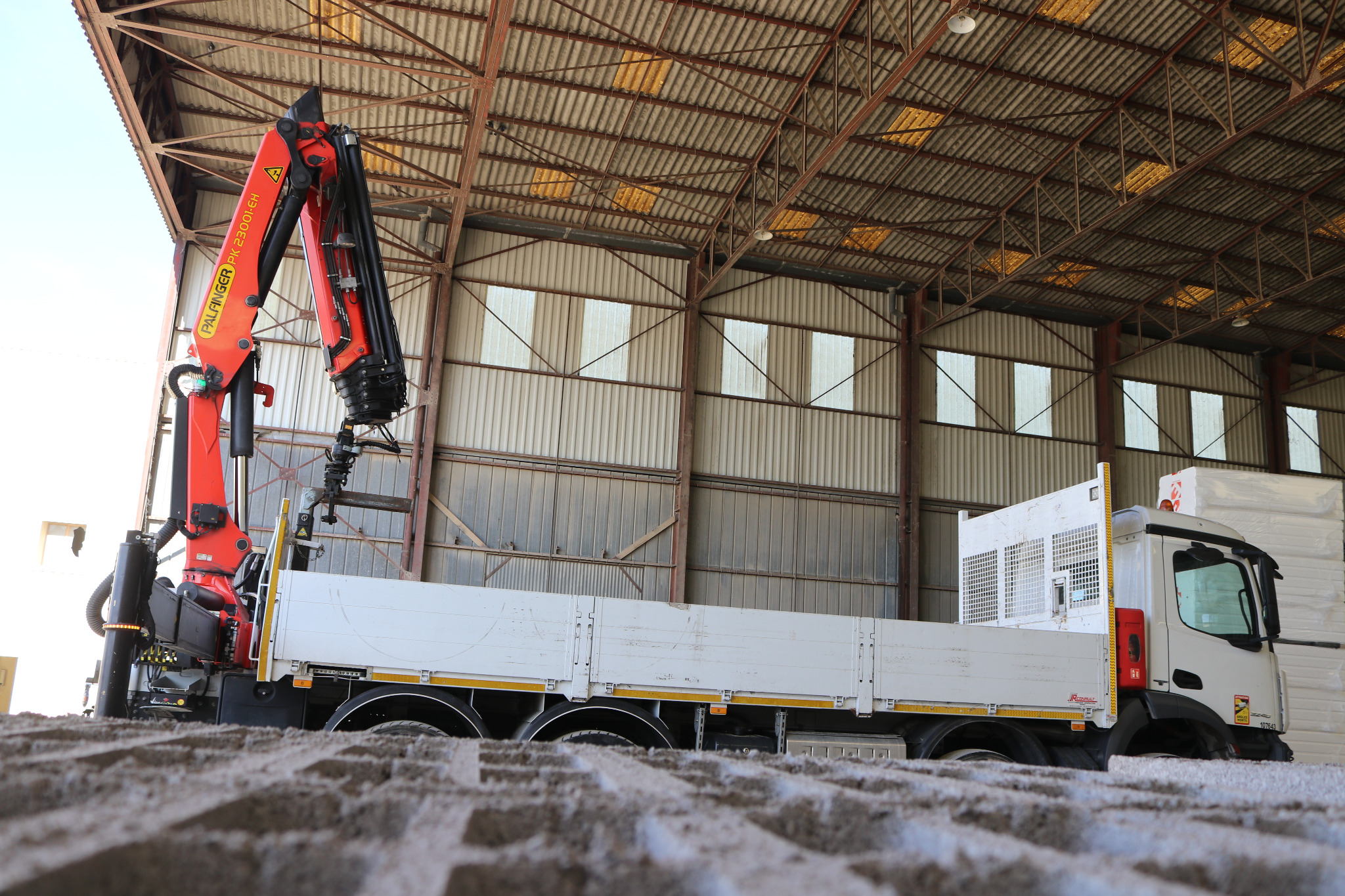 Ciężarówka z zamontowanym dźwigiem Palfinger PK 23001-EH na tle magazynu z metalową konstrukcją dachu i oknami, widziana z poziomu gruntu.