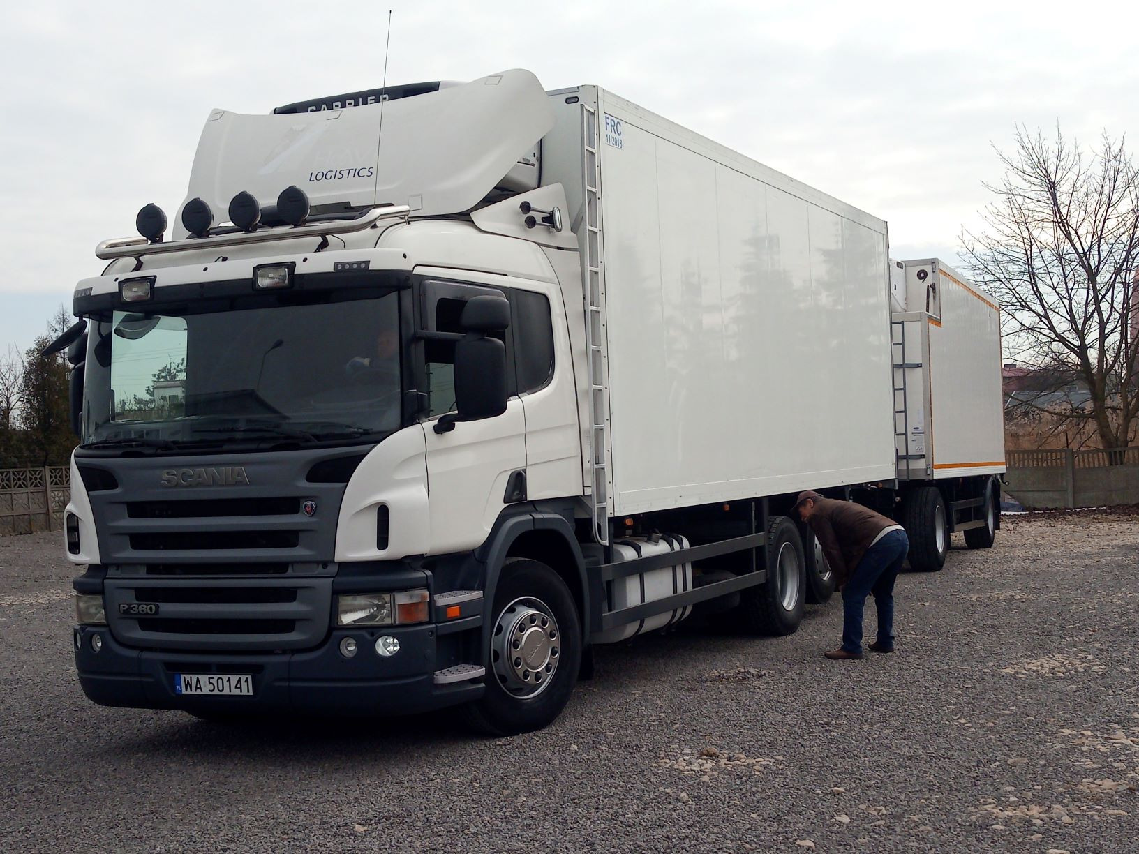 Biała ciężarówka chłodnia Scania P360 z naczepą, mężczyzna sprawdza oponę na żwirowym parkingu.