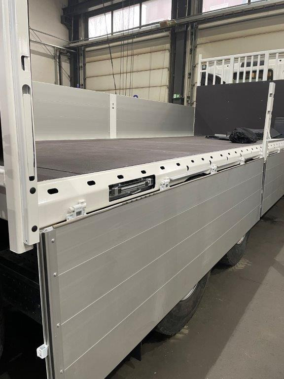 Wnętrze skrzyni ładunkowej nowego, białego samochodu ciężarowego z aluminiowymi burtami bocznymi, widoczne mocowania i czarna podłoga, w tle hala produkcyjna.