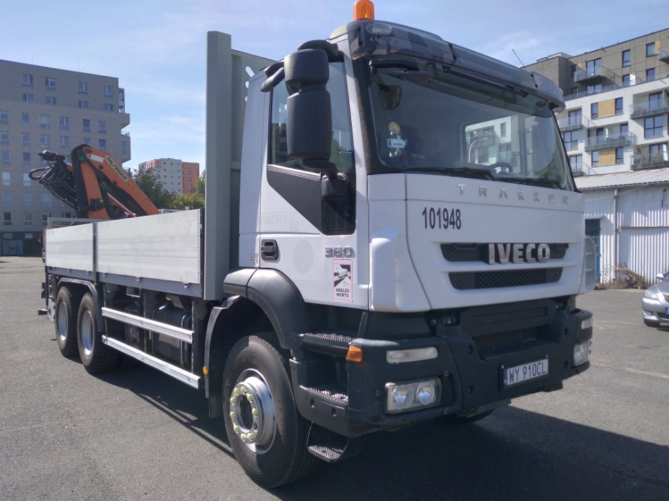 Biała ciężarówka Iveco Trakker z zamontowanym pomarańczowym dźwigiem Palfinger na platformie, zaparkowana na placu budowy z widokiem na nowoczesne budynki mieszkalne w tle.