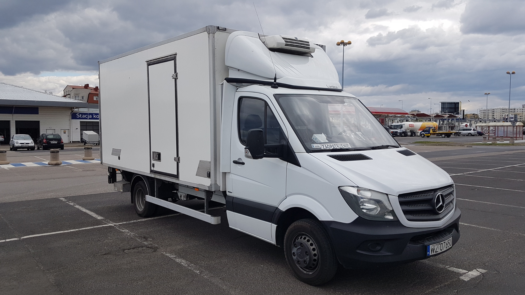 Biały samochód dostawczy typu chłodnia marki Mercedes-Benz zaparkowany na parkingu z widocznym numerem rejestracyjnym i napisem 'ZAOPATRZENIE' za szybą.