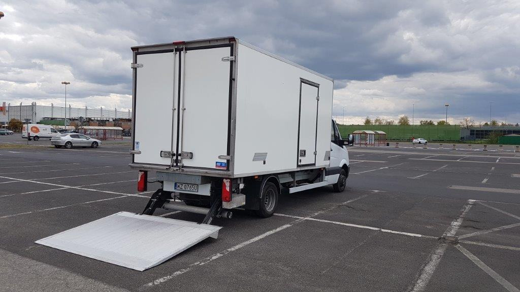 Tył białego samochodu dostawczego z podniesioną rampą załadowczą na pustym parkingu, widoczne tablice rejestracyjne.