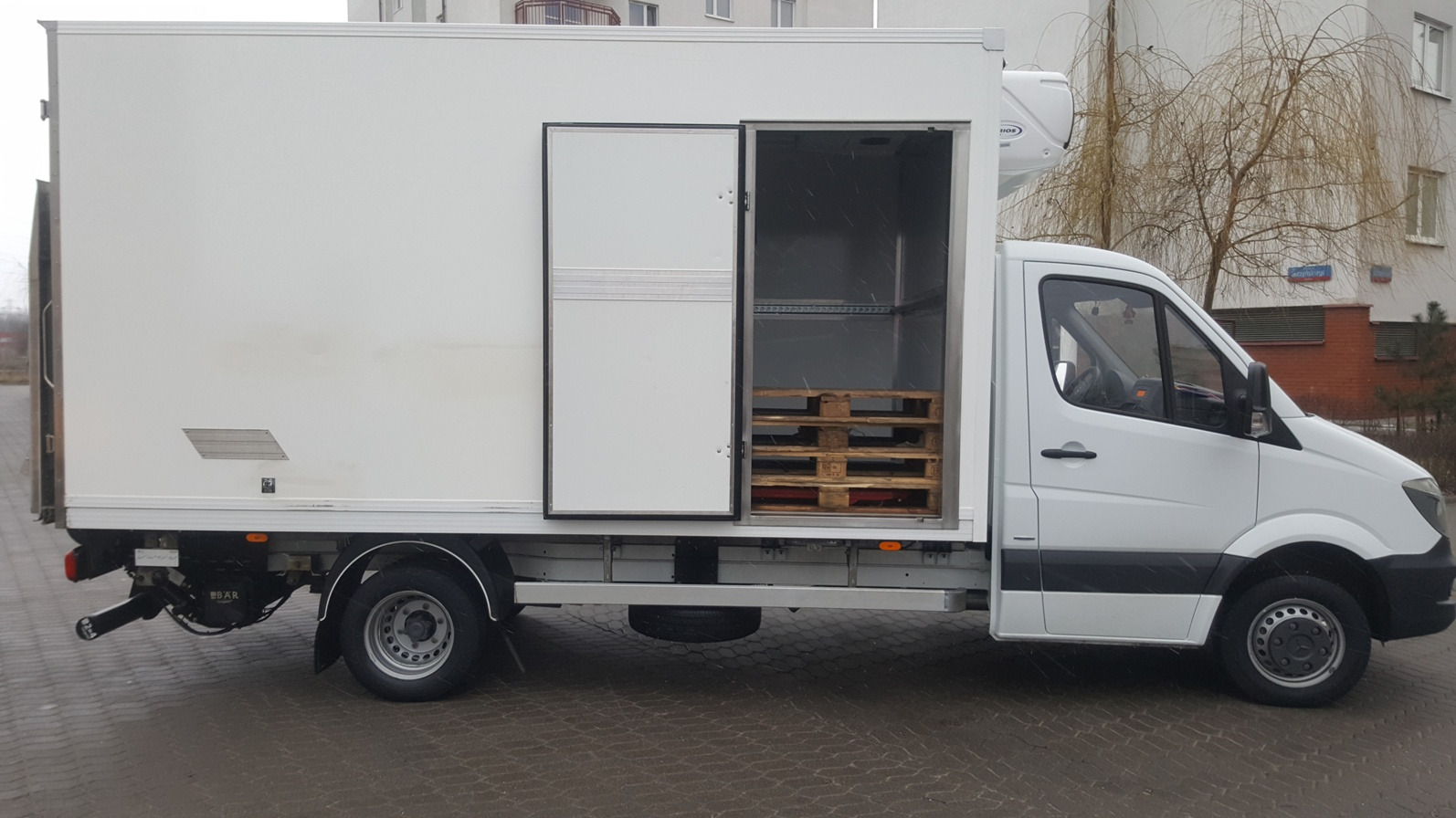 Biały samochód dostawczy z otwartymi drzwiami przestrzeni ładunkowej, w której widoczne są trzy drewniane palety.