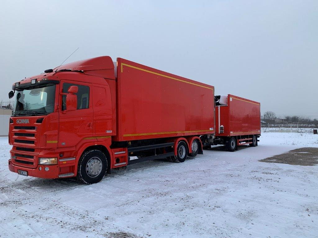 Czerwony ciągnik siodłowy Scania z dwiema naczepami na zaśnieżonym placu, widok z boku, pochmurne niebo.