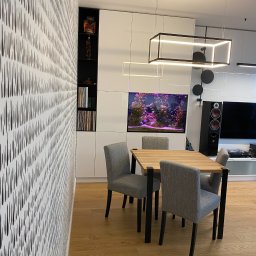 EMKA Marczak Łukasz - Nowoczesna jadalnia z drewnianym stołem, szarymi krzesłami i akwarium w zabudowie meblowej, oświetlona geometryczną lampą wiszącą.