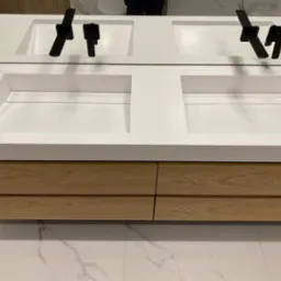 Nowoczesna łazienka z podwójną, wpuszczaną umywalką z białego konglomeratu, czarną armaturą i szafką z jasnego drewna na tle marmurowej podłogi.