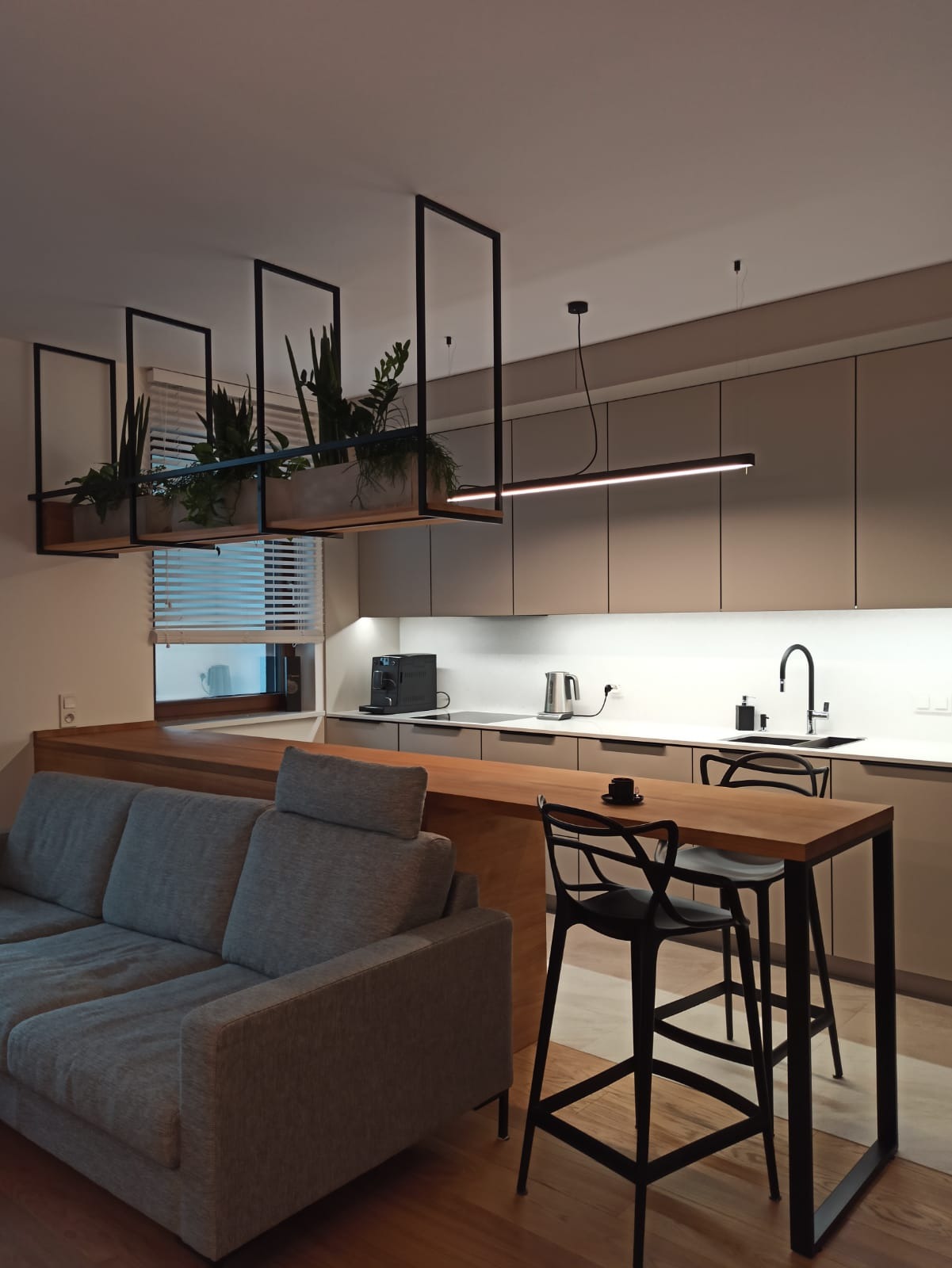 Nowoczesna kuchnia w odcieniach beżu z drewnianym blatem, czarnymi hokerami i wiszącą konstrukcją z roślinami doniczkowymi, oświetlona listwą LED.