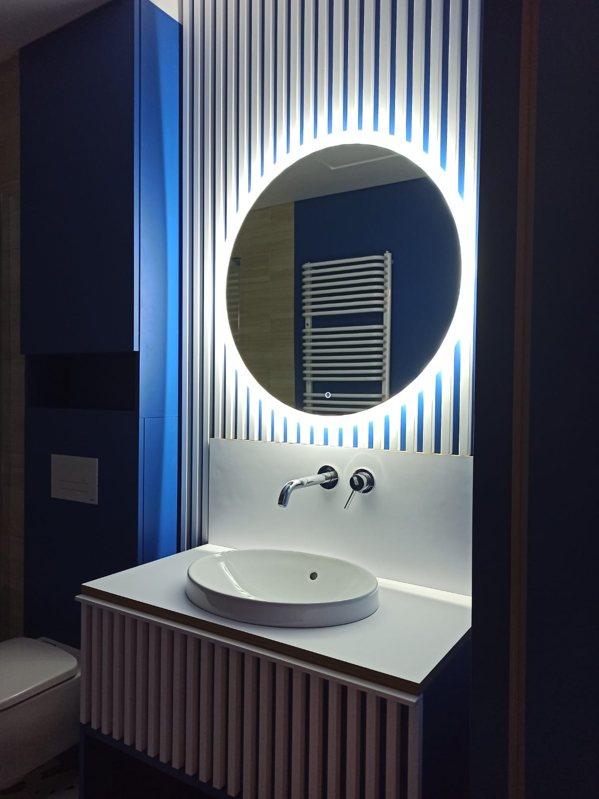 Nowoczesna łazienka z umywalką nablatową, okrągłym lustrem z podświetleniem LED, niebieskimi akcentami i grzejnikiem drabinkowym w tle odbicia.