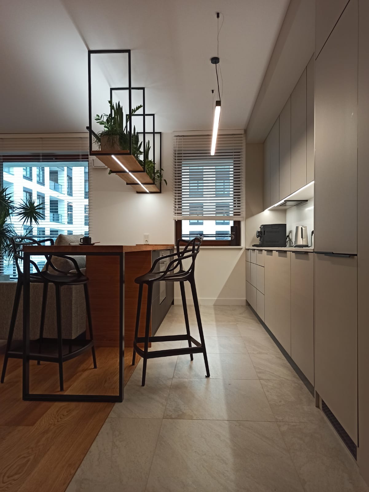 Nowoczesna kuchnia z wyspą i hokerami, oświetleniem LED i wiszącą konstrukcją z roślinami, widok na miasto przez okno.