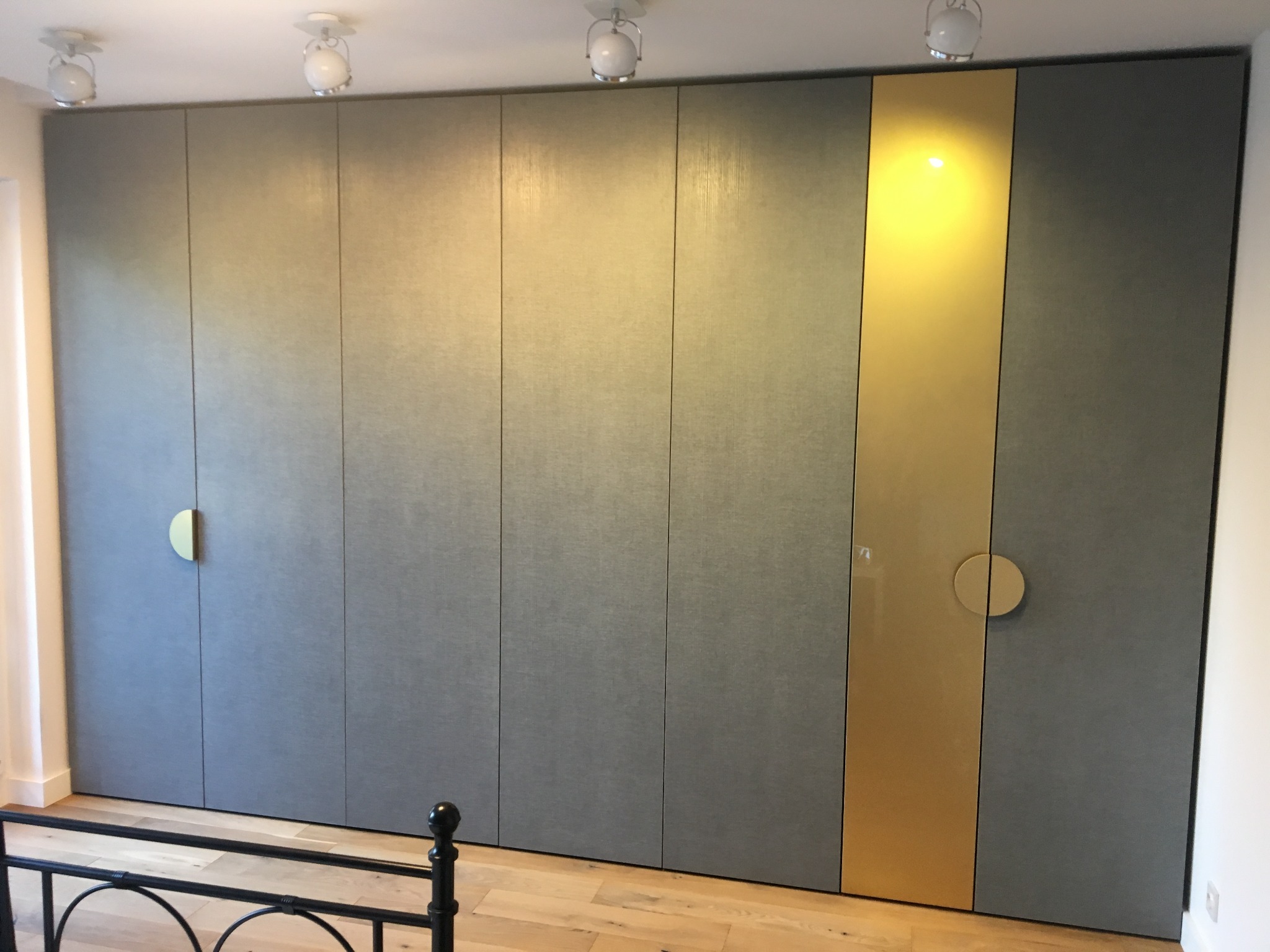 Duża szafa wnękowa z szarymi frontami i złotym pionowym panelem, okrągłe uchwyty, widoczny fragment łóżka.