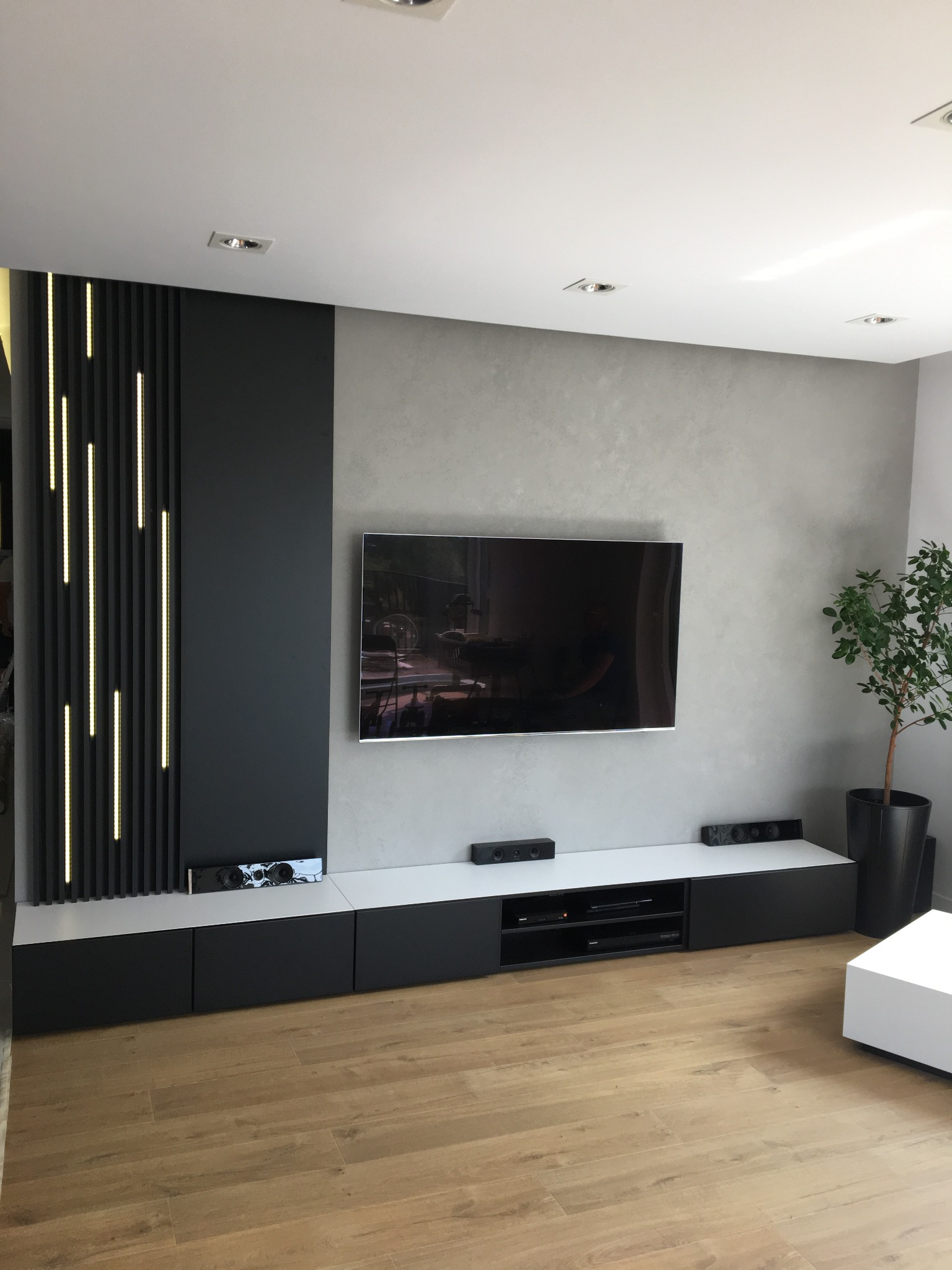 Nowoczesna ściana telewizyjna z podświetlanymi panelami pionowymi, szafkami RTV w kolorze czarno-białym i dekoracyjnym tynkiem na ścianie w salonie.